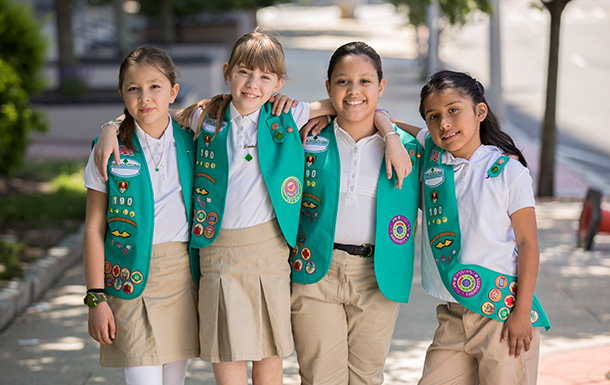 Four young Girl Scouts hug while they smile at the camera. Girl Scouts helps girls transform into women as they learn new life skills. 