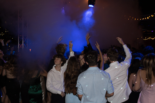 With their hands raised, a group of friends dance to the music. The fog machine produces fog that engulfs the crowd, making for an even more awe-inspiring setting. 