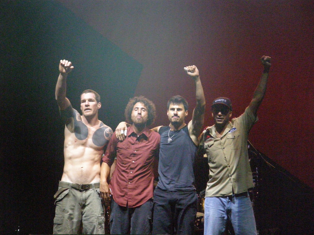Rage Against the Machine (RATM) members pose at their Vegoose 2007 performance. From left to right, Tim Commerford (bassist), Zack De La Rocha (vocalist), Brad Wilk (drummer) and Tom Morello (guitarist) stand with their fists in the air.  Rage Against The Machine @ Vegoose 2007  by  Scott Penner  is licensed under  CC BY-SA 2.0 DEED. 