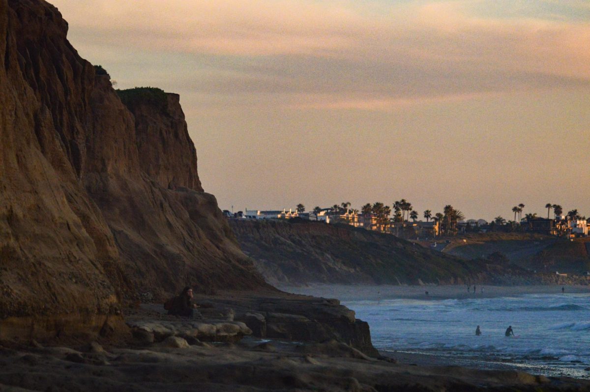 A woman sits atop a rock platform and overlooks the beach. Near the Carlsbad Tide Pools, surfers float in the shallow end, making their way into deeper waters. 
