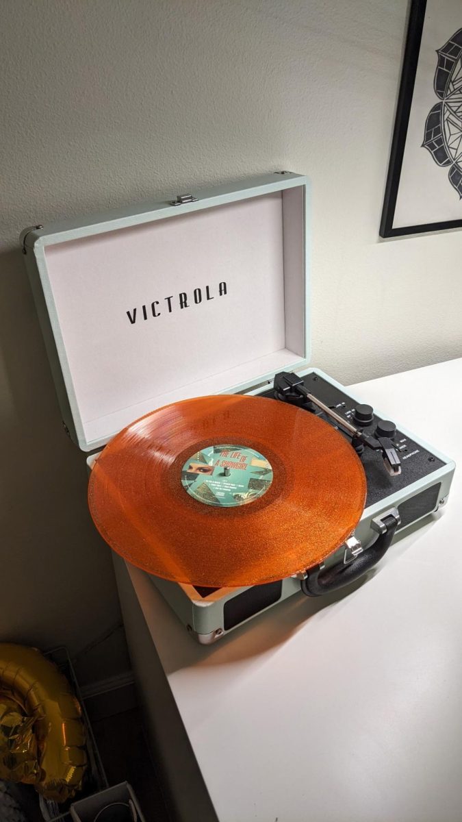 A Taylor Swift vinyl sits on a vinyl player behind a plain white wall. The vinyl is a sparkly orange, with the title of the album “The Life of a Showgirl” in the middle. 
