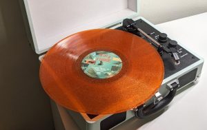 A Taylor Swift vinyl sits on a vinyl player behind a plain white wall. The vinyl is a sparkly orange, with the title of the album “The Life of a Showgirl” in the middle. 
